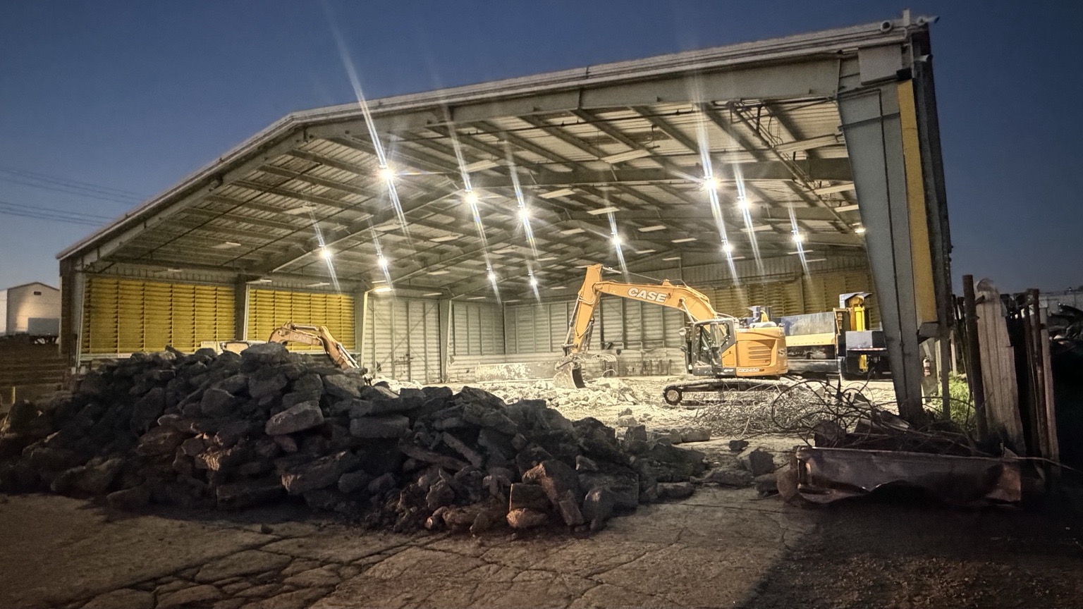 Excavators demolishing a concrete slab inside a large shop at dusk